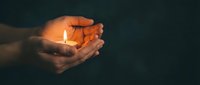 Close-up of hands holding a lit candle against dark background, symbolizing hope, spirituality, and warmth for memorial or inspirational concepts.