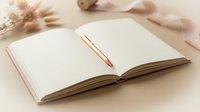Open notebook with blank pages and pen on neutral beige surface, surrounded by dried flowers and ribbon, ready for writing, journaling, planning, or creative projects.