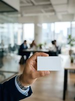Man in business attire holding blank white card in modern office environment with colleagues meeting in background, suitable for corporate branding and networking concepts.