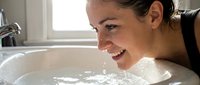 Woman with dark hair and light skin tone enjoying a relaxing soak in a bathtub filled with warm water and bubbles, smiling contentedly during self-care and wellness routine at home.