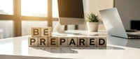 Wooden blocks spelling "BE PREPARED" on a modern office desk with computer monitor, laptop, and potted plant in bright workspace environment.