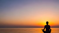 Silhouette of person meditating in lotus pose at sunset over calm ocean water with colorful sky gradient from purple to orange for wellness and mindfulness.