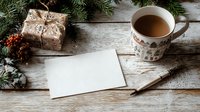 Cozy winter scene with hot coffee, blank card, and wrapped gift on rustic wooden table surrounded by pine branches and festive decorations.