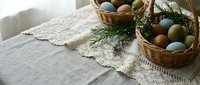 Easter eggs in wicker baskets with evergreen branches on linen tablecloth with lace trim for spring holiday celebration and seasonal home decor.