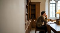 Middle-aged Caucasian man in contemplative pose at wooden desk in traditional library study with built-in bookshelves and arched window for academic workspace.