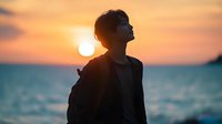 Young Asian man silhouette against dramatic orange sunset sky and ocean backdrop, wearing casual jacket, face turned up towards warm sunlight in peaceful contemplative moment.