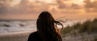 Young woman with long flowing hair watching sunset over ocean waves from beach, peaceful moment of solitude and reflection during golden hour.