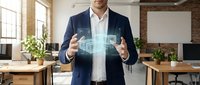 Mature male professional in navy blazer and white shirt gestures toward glowing digital hologram interface in modern office workspace with brick walls and natural lighting.