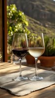 Red and white wine glasses on rustic wooden table with mountain backdrop, sunlit terrace setting creates romantic atmosphere for wine tasting experience.