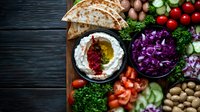 Fresh Mediterranean mezze platter with labneh dip, spices, flatbread, chopped vegetables, and legumes on dark wooden background, overhead view of colorful appetizers.