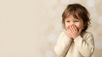Adorable toddler boy with brown hair covering mouth while laughing, wearing cream sweater against soft bokeh background for family content.