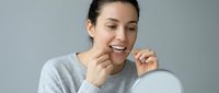 Young woman with dark hair and fair complexion flossing teeth while looking in mirror, promoting dental hygiene and oral care routine.