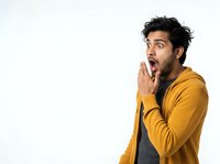 Young man with dark hair and South Asian ethnicity wearing a mustard yellow cardigan over gray shirt, expressing shock and surprise with hand near mouth against white background. Suitable for advertising, marketing, emotional reactions, and lifestyle content.