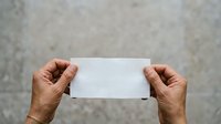 Middle-aged man with medium skin tone holding blank white card or paper against neutral gray concrete wall background for mockup and branding purposes.