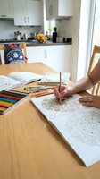 Adult woman coloring intricate mandala patterns at wooden dining table with colored pencils, sketchbooks, and art supplies in bright modern kitchen setting.