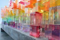 Colorful translucent acrylic blocks arranged in rows on concrete surface, creating vibrant rainbow effect with light passing through geometric shapes.