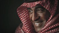 Middle-aged Middle Eastern man wearing traditional red and white keffiyeh headdress smiling against dark background, conveying warmth and cultural heritage.