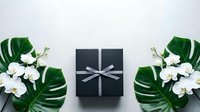Black gift box with silver ribbon surrounded by white orchids and tropical monstera leaves on white background for special occasions and celebrations.
