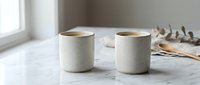 Two ceramic mugs filled with coffee sit on a marble countertop near a window, styled with dried botanicals and natural decor for home and lifestyle use.