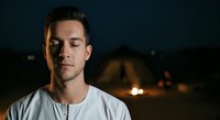 Young man with dark hair and facial stubble wearing casual white shirt, looking contemplative against blurred nighttime background with warm lights, suitable for portrait and lifestyle content.