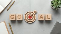 Wooden blocks spelling goal with target bullseye icon on concrete desk, surrounded by office supplies and succulent plant for business motivation.