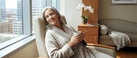 Relaxed mature woman enjoying coffee in modern apartment living room with city view through large windows.