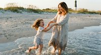 Mother and daughter holding hands walking on beach at sunset with lighthouse in background during family vacation.