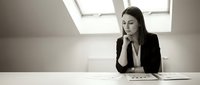 Professional businesswoman in contemplative pose at modern office desk with documents under natural skylight illumination for corporate presentations.