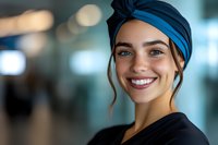 Young female healthcare worker wearing blue surgical cap smiles warmly at camera. Natural lighting and blurred hospital background create professional medical setting.