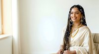 Young woman with dark skin tone and long braided hair wearing a cream-colored satin shirt, smiling at camera in bright indoor setting. Portrait for lifestyle, fashion, and professional use.