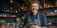 Confident mature male cafe owner with gray beard using digital tablet in stylish coffee shop with wooden shelves and warm lighting.