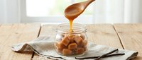 Wooden spoon drizzling golden caramel sauce over nuts in glass jar on rustic kitchen table. Sweet dessert topping preparation for baking and cooking.