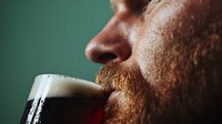 Close up profile of bearded man drinking dark craft beer with thick creamy foam head against green background. Macro shot captures rich texture and bubbles.