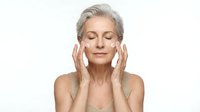 Mature woman applying facial cream with closed eyes showing skincare routine for anti-aging beauty treatment on white background.