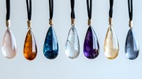 Colorful crystal pendants on black cords arranged in row showing variety of natural gemstone drops including rose quartz, amber, blue topaz, clear quartz, amethyst and citrine.