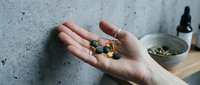 Caucasian hand holding assorted supplement capsules and pills against concrete wall, with bowl of seeds and dropper bottle nearby for wellness and nutrition content.