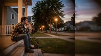 Young man sitting on porch steps in suburban neighborhood at dusk, contemplating life decisions while enjoying peaceful evening atmosphere in residential area.