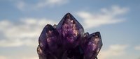 Stunning deep purple amethyst crystal cluster against a cloudy sky, showcasing natural gemstone formation with rich violet hues and sharp facets.