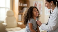 Female pediatrician examining happy African American girl with stethoscope in modern medical office during routine checkup appointment.