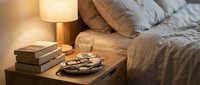 Cozy bedroom nightstand with warm lamp light, books, glasses and coffee cup creating peaceful evening atmosphere for relaxation and reading.