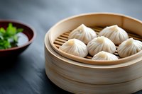 Steamed dumplings in traditional bamboo steamer with fresh herbs and dipping sauce on dark background, selective focus on Asian cuisine delicacy.