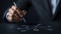Professional in dark suit drawing business network diagram or mind map on transparent surface, strategic planning concept in low key lighting.