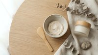 Overhead view of natural skincare products with wooden applicator on light wood surface, cream jar, and dried eucalyptus branches for cosmetics, beauty, wellness, and spa design projects.