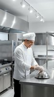 Professional male chef in white uniform and hat preparing food in modern commercial kitchen with stainless steel equipment and professional lighting.
