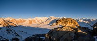 Majestic snow-covered mountain range at sunrise with golden peaks, deep valleys, and clear blue sky, suitable for travel and nature backgrounds.