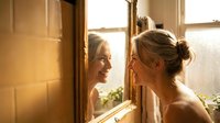 Young blonde woman applying skincare routine while looking in bathroom mirror during golden hour morning light creating warm glow for wellness content.