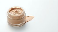 Open glass jar with creamy beige foundation makeup and smear sample on white background. Cosmetic product for skincare and beauty routine applications.