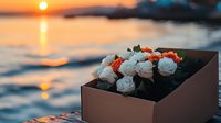 Romantic bouquet of white and orange roses in gift box against stunning ocean sunset background, golden hour reflection on water creates magical atmosphere.