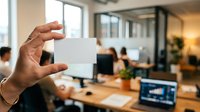 Professional holding blank white business card in modern office environment with colleagues working at desks in background, suitable for corporate branding and business networking.
