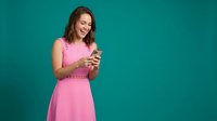 Young Caucasian woman in pink dress smiling while using smartphone on turquoise background for social media marketing and communication concepts.